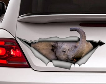 May include: A gray baby elephant with its trunk raised is peeking out of a ripped hole in a white car. The elephant's eye is visible, and its trunk is curled.