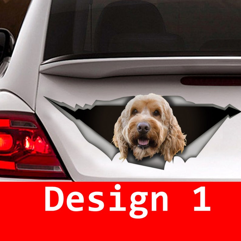 May include: A brown and white dog's head peeking out of a ripped car window decal. The decal is white with a black outline and the dog is looking directly at the camera. The text "Design 1" is below the decal.