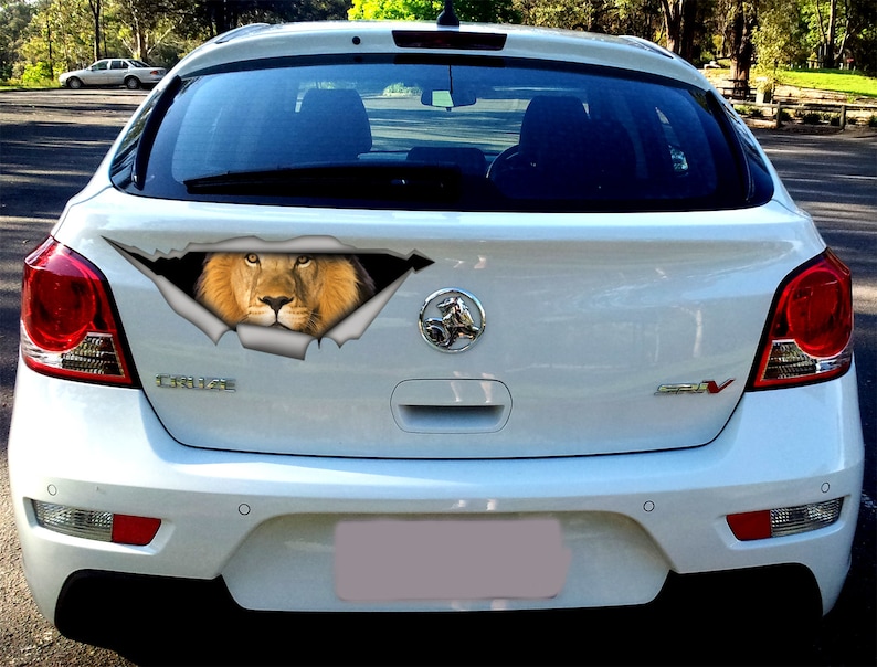 May include: A white car with a lion's head peeking out of a ripped hole in the back window. The lion has a brown mane and a serious expression.