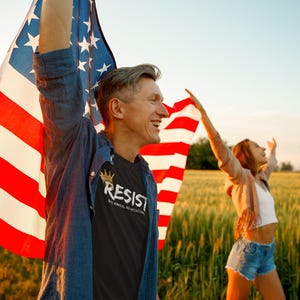 May include: Two people holding American flags in a field. The person on the left is wearing a black t-shirt with the words "Resist" and a crown printed on it.