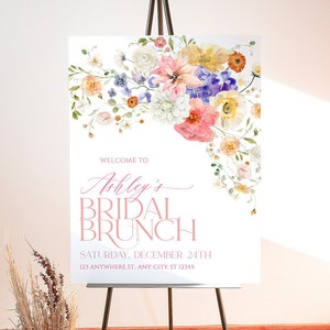 May include: A white sign with a floral design in shades of pink, yellow, blue, and green. The text on the sign reads "Welcome to Ashley's Bridal Brunch, Saturday, December 24th, 123 Anywhere St., Any City, ST 12345".