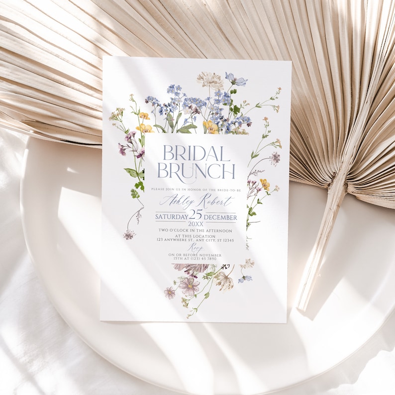 May include: A white invitation card with a floral border. The text reads "Bridal Brunch" in blue and white lettering. The invitation is for Ashley Robert's bridal brunch on Saturday, December 25th, 20XX at 2:00 PM. The location is 123 Anywhere St., Any City, ST 12345. RSVP by November 19th at (123) 45 7890.