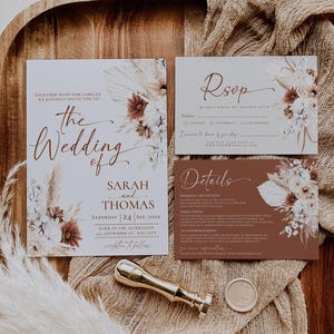 May include: A wedding invitation suite featuring a main invitation, an RSVP card, and a details card, all decorated with dried floral illustrations in white, cream, and rust tones. A gold wax seal stamp and a circular wax seal are included.