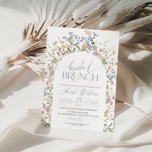 Pu&ograve; includere: Un invito per un brunch nuziale con la scritta "bridal brunch" in elegante corsivo. L'invito include la data, l'ora e il luogo, circondati da un delicato bordo floreale con fiori blu, viola e gialli.
