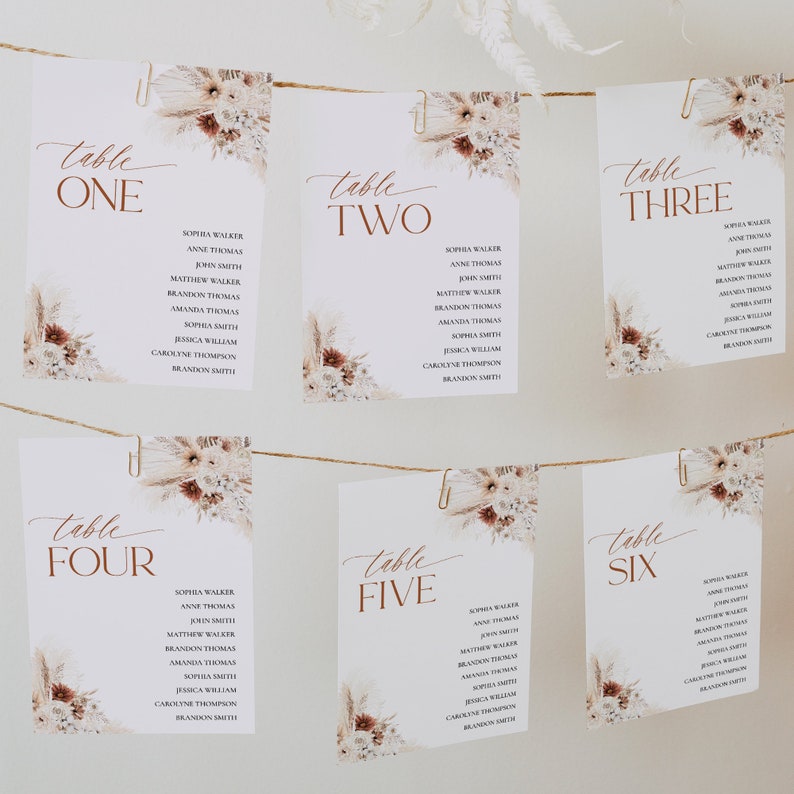 Puede incluir: Seis tarjetas de mesa blancas con n&uacute;meros de mesa en escritura marr&oacute;n y nombres de invitados, decoradas con arreglos florales. Las tarjetas est&aacute;n suspendidas por clips en una cuerda, adecuadas para bodas o eventos.