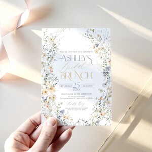 May include: A white floral bridal brunch invitation with gold lettering. The invitation reads "You're invited to celebrate Ashley's Bridal Brunch, Saturday 25 August 20XX, Two o'clock in the afternoon at this location 123 Anywhere St. Any City, ST 12345, RSVP on or before November 15th at (123) 45 7890".