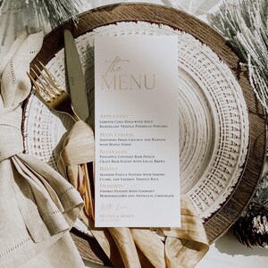 May include: A vertical white menu card with gold and dark text reading The Menu, Appetizers, Main Course, Beverages, Buffet, Desserts, and Olivia & Robin. It rests on a white plate with a gold fork, knife, and beige linen napkin.