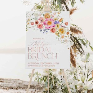 May include: A white rectangular sign on a wooden easel featuring a watercolor floral arrangement in pink, orange, and yellow. The text reads: WELCOME TO Ashley's BRIDAL BRUNCH SATURDAY, DECEMBER 24TH 123 ANYWHERE ST. ANY CITY, ST 12345.