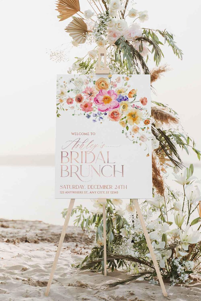 May include: A white rectangular sign on a wooden easel featuring a watercolor floral arrangement in pink, orange, and yellow. The text reads: WELCOME TO Ashley's BRIDAL BRUNCH SATURDAY, DECEMBER 24TH 123 ANYWHERE ST. ANY CITY, ST 12345.