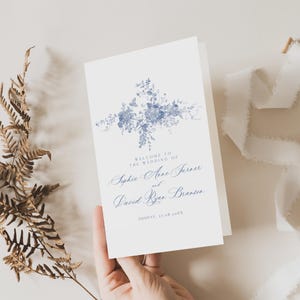 May include: A white wedding program with a blue floral design and text. The program reads "Welcome to the Wedding of Sophie Anne Turner and David Ryan Branson" with the date below. A hand holds the program.