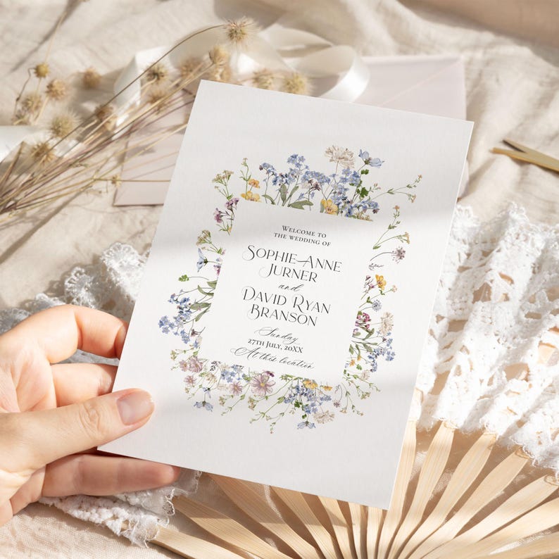 May include: Wedding ceremony program with a floral border of blue, yellow, and pink wildflowers. The text reads "Welcome to the wedding of Sophie Anne Jurner and David Ryan Branson, Sunday, 27th July, 20XX, At the beatin"