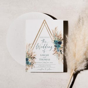 Puede incluir: Una invitaci&oacute;n de boda con un dise&ntilde;o geom&eacute;trico dorado y detalles florales. La invitaci&oacute;n dice "The Wedding of Sarah and Thomas" con la fecha y la hora. Una pluma de hierba de la Pampa beige y esponjosa est&aacute; en el fondo.