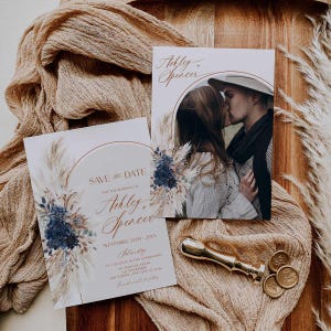 May include: Wedding invitation suite featuring a photo of a couple kissing, and a "Save the Date" card with floral and pampas grass accents. The cards have the names "Ashley & Spencer" in elegant script. A gold wax seal and stamps are also present.