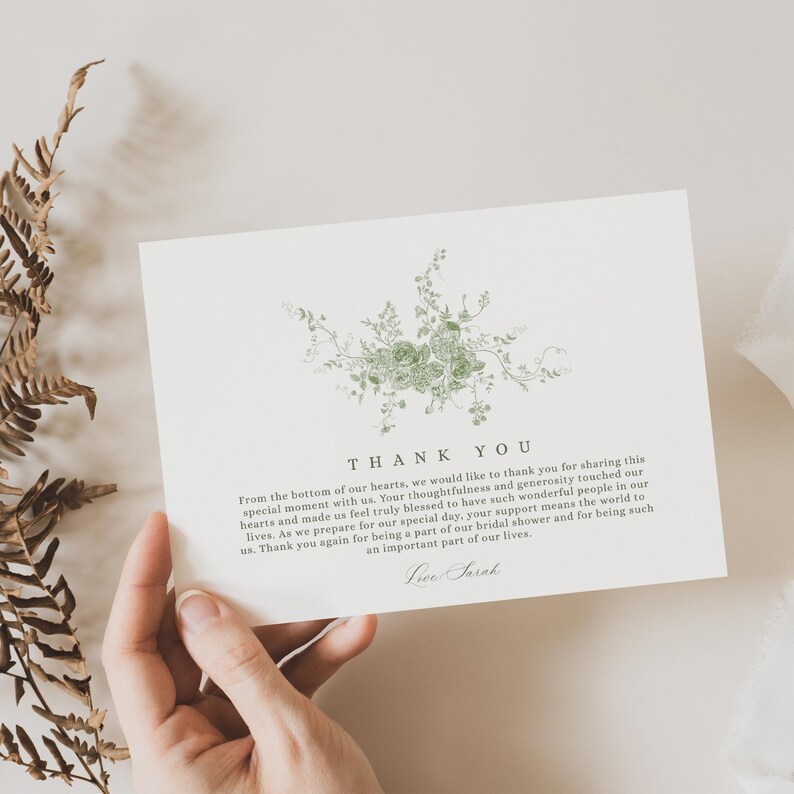 May include: A cream-colored thank you card with a green floral design. The card has the words "THANK YOU" at the top, with a message below. The card is held by a person.