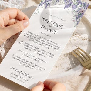 May include: A rectangular white wedding welcome card featuring a purple floral illustration at the top. The text reads: TO OUR FAMILY & FRIENDS, WELCOME and THANKS, Dear guests, followed by a message, with heartfelt thanks, SOPHIE & DAVID, 27.08.20xx.