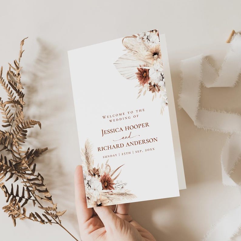 May include: A white wedding program with a floral design featuring dried pampas grass, white and red flowers. The text reads "Welcome to the wedding of Jessica Hooper and Richard Anderson, Sunday 25th Sep, 20XX".
