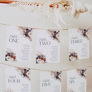 May include: Six white rectangular wedding seating charts hanging on twine with gold clips. Each card features navy blue and burgundy floral accents, pampas grass, and the text Table One through Table Six with lists of names printed in dark blue ink.