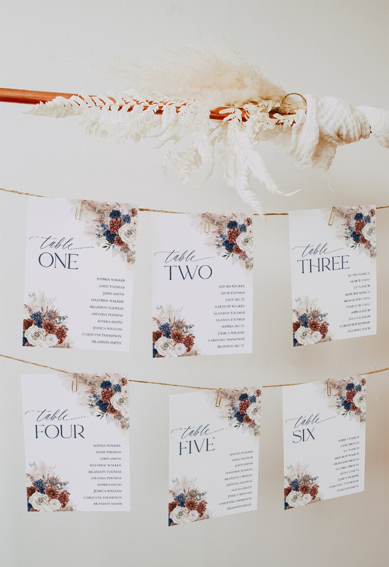 May include: Six white rectangular wedding seating charts hanging on twine with gold clips. Each card features navy blue and burgundy floral accents, pampas grass, and the text Table One through Table Six with lists of names printed in dark blue ink.