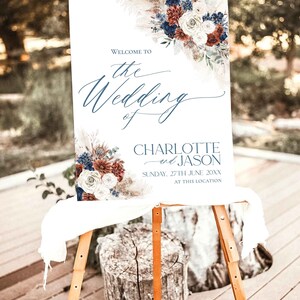 May include: A white wedding sign with a floral design in shades of blue, red, and white. The sign reads "Welcome to the Wedding of Charlotte and Jason, Sunday, 27th June 20XX, at this location."
