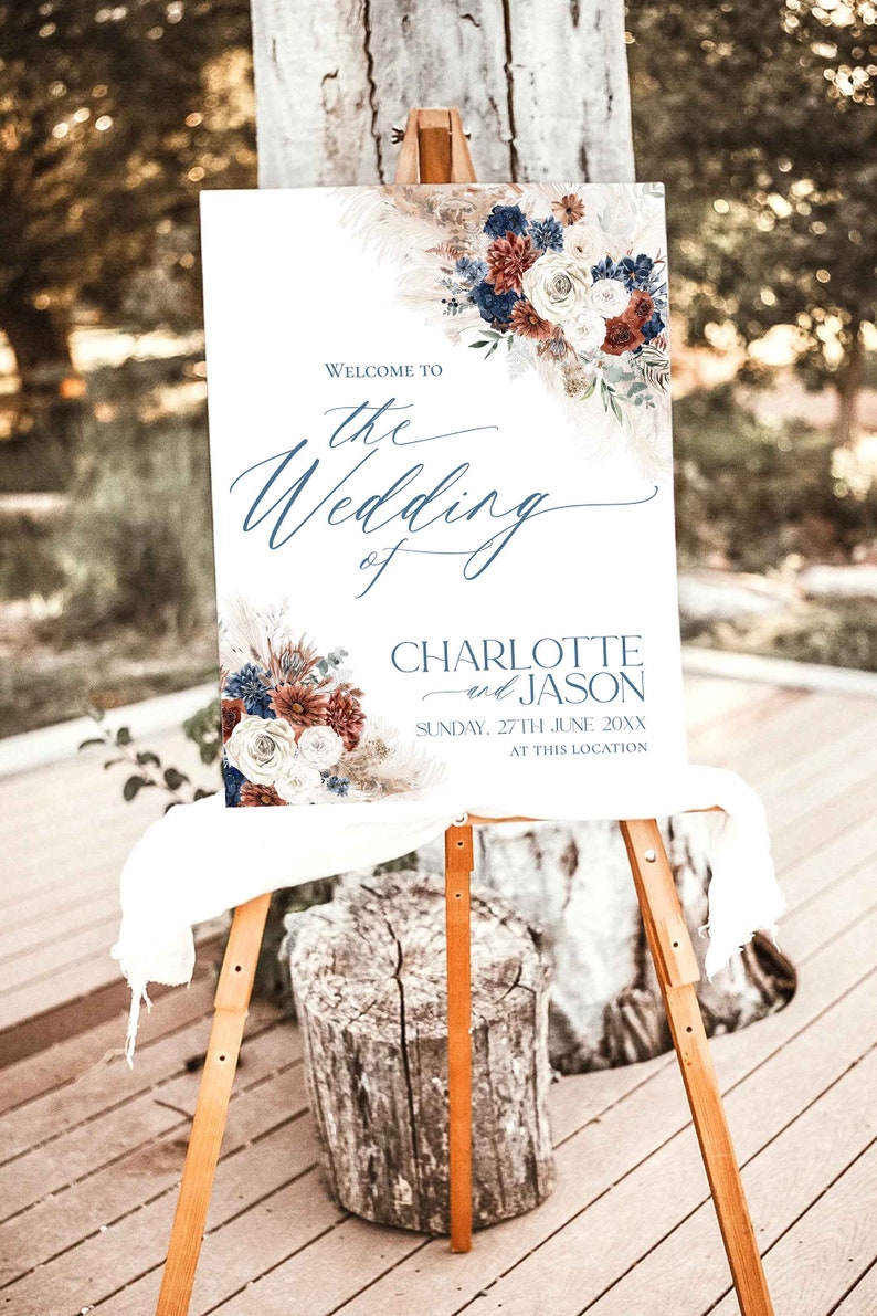 May include: A white wedding sign with a floral design in shades of blue, red, and white. The sign reads "Welcome to the Wedding of Charlotte and Jason, Sunday, 27th June 20XX, at this location."