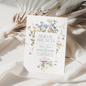 May include: A white bridal brunch invitation with a floral design featuring blue and yellow flowers. The text reads "Bridal Brunch" in a blue font and "Ashley Robert" in a cursive font. The date is Saturday, December 25th, 20XX. The time is two o'clock in the afternoon. The location is 123 Anywhere St., Any City, ST 12345. The RSVP date is on or before November 15th at 0231 45 2890.