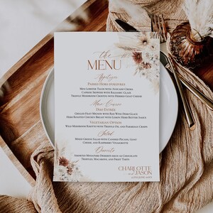 May include: A rectangular wedding menu card featuring the text "the MENU" and "CHARLOTTE and JASON 27TH JUNE 20XX" with printed floral illustrations of dried pampas grass, white roses, and rust-colored flowers in the top right and bottom left corners.