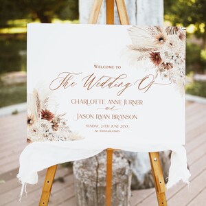 May include: A rectangular white wedding welcome sign on a wooden easel, featuring brown script text reading "Welcome to The Wedding Of Charlotte Anne Jurner and Jason Ryan Branson, Sunday, 24th June 20XX, At This Location" with dried floral accents.