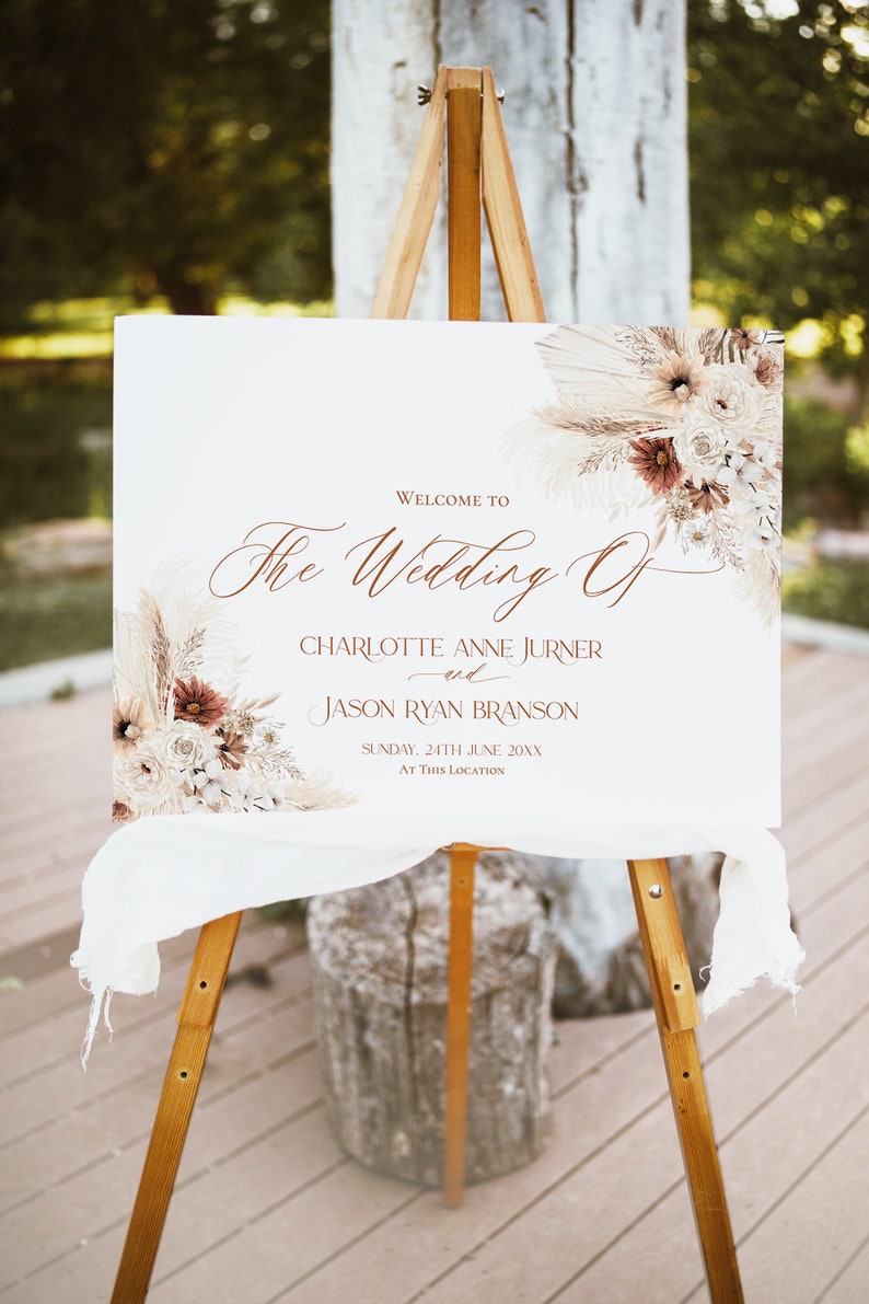 May include: A rectangular white wedding welcome sign on a wooden easel, featuring brown script text reading "Welcome to The Wedding Of Charlotte Anne Jurner and Jason Ryan Branson, Sunday, 24th June 20XX, At This Location" with dried floral accents.