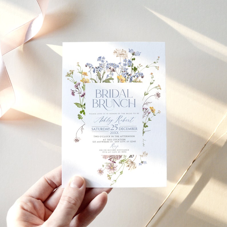 May include: A white invitation card with a floral border. The text reads "Bridal Brunch" in a blue font, followed by "Please join us in honor of the bride to be Ashley Robert Saturday 25 December 20XX Two o'clock in the afternoon at this location 123 Anywhere St. Any City, ST 12345 RSVP on or before November 15th at (123) 45 7890"