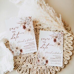 May include: Two save the date cards with a floral design and the text "save the date" in a cursive font. The cards are on a woven placemat with a dried flower arrangement.