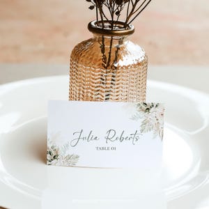 May include: White place card with a floral design and the text "Julia Roberts Table 01". The place card is sitting on a white plate with a small glass vase with dried flowers.