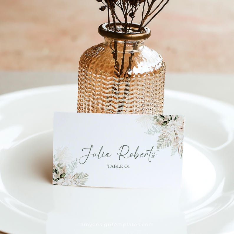 May include: White place card with a floral design and the text "Julia Roberts Table 01". The place card is sitting on a white plate with a small glass vase with dried flowers.