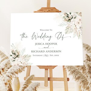 May include: A white rectangular wedding welcome sign on a wooden easel, featuring floral arrangements of pampas grass and green leaves in the corners. The text reads: WELCOME TO the Wedding Of JESICA HOOPER and RICHARD ANDERSON SATURDAY | 24 | SEP, 20XX.