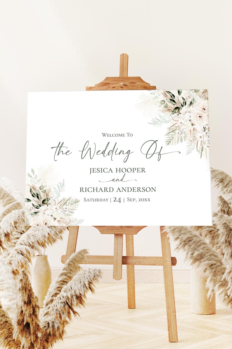 May include: A white rectangular wedding welcome sign on a wooden easel, featuring floral arrangements of pampas grass and green leaves in the corners. The text reads: WELCOME TO the Wedding Of JESICA HOOPER and RICHARD ANDERSON SATURDAY | 24 | SEP, 20XX.