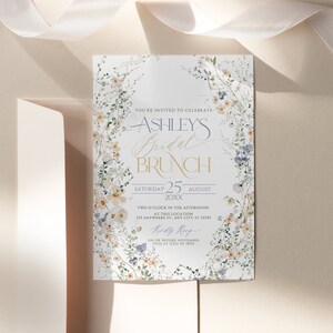 May include: A floral bridal brunch invitation with a white background and a wreath of watercolor flowers. The text reads "You're invited to celebrate Ashley's Bridal Brunch, Saturday 25 August 20XX, Two o'clock in the afternoon at this location: 123 Anywhere St. Any City, ST 12345, RSVP on or before November 15th at (123) 45 7890."
