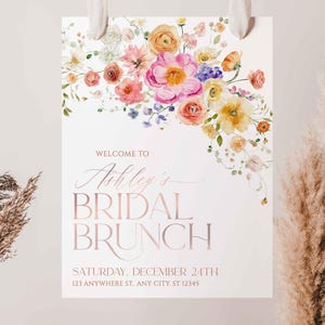 May include: A white rectangular sign featuring a watercolor floral arrangement in shades of pink, yellow, and orange at the top. The text reads: WELCOME TO Ashley's BRIDAL BRUNCH SATURDAY, DECEMBER 24TH 123 ANYWHERE ST, ANY CITY, ST 12345.