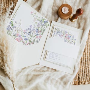 K&ouml;nnte beinhalten: Ein wei&szlig;es Einladungsset mit einem floralen Umschlagfutter mit rosa, blauen und gr&uuml;nen Wildblumen. Das Set enth&auml;lt eine Karte mit einer wei&szlig;en Papierbanderole, einen h&ouml;lzernen Wachssiegelstempel mit botanischem Motiv und einen getrockneten Pampasgrasst&auml;ngel.