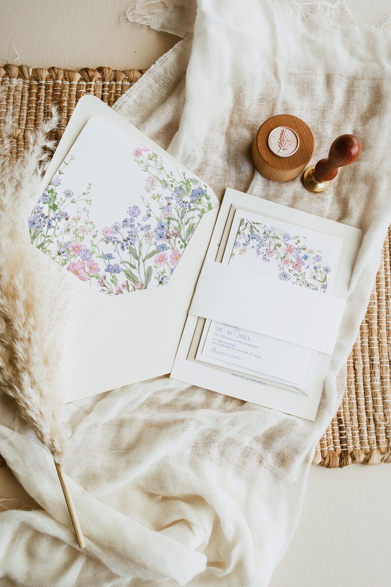 K&ouml;nnte beinhalten: Ein wei&szlig;es Einladungsset mit einem floralen Umschlagfutter mit rosa, blauen und gr&uuml;nen Wildblumen. Das Set enth&auml;lt eine Karte mit einer wei&szlig;en Papierbanderole, einen h&ouml;lzernen Wachssiegelstempel mit botanischem Motiv und einen getrockneten Pampasgrasst&auml;ngel.