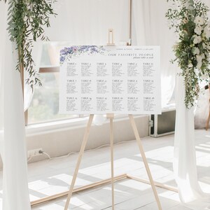 Puede incluir: Un plano de asientos de boda con un dise&ntilde;o floral en tonos morados y azules. El plano est&aacute; en un caballete y enumera los n&uacute;meros de mesa del 1 al 18. El texto en la parte superior dice "Nuestra Gente Favorita, Por Favor Tomen Asiento."