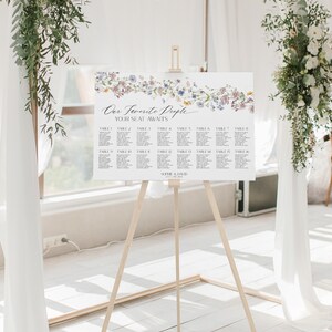 May include: A wedding seating chart with a floral design. The chart is on an easel and has the text "Our Favorite People Your Seat Awaits" at the top. The chart lists tables 1 through 16 with names under each table number.