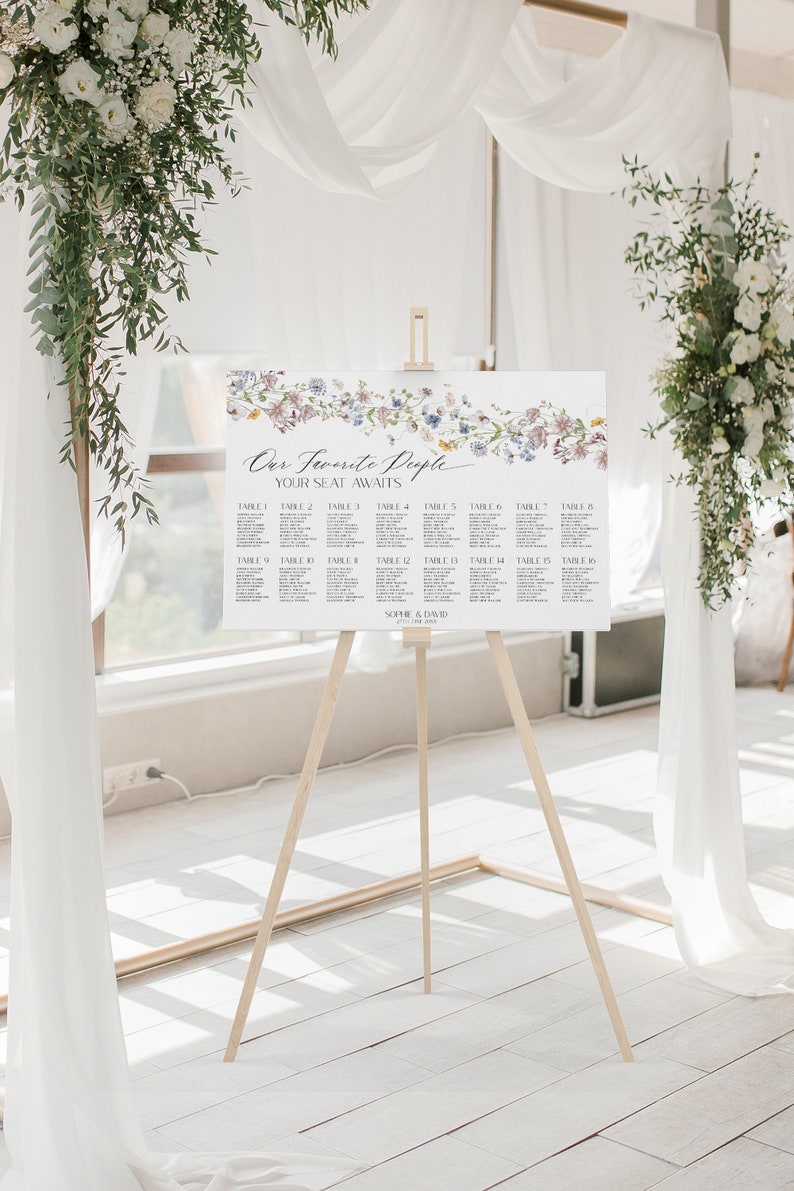 May include: A wedding seating chart with a floral design. The chart is on an easel and has the text "Our Favorite People Your Seat Awaits" at the top. The chart lists tables 1 through 16 with names under each table number.
