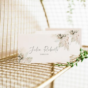 May include: A white place card with a floral design in shades of green and pink. The text "Julia Roberts" and "Table 01" is printed in gray.