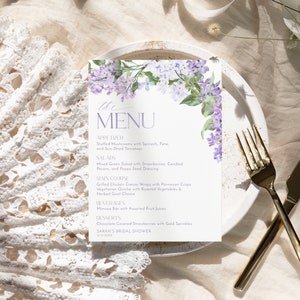 Op de afbeelding: Een lila bloemenmenukaart met de tekst "the MENU" in lichtpaarse lettertype. Het menu bevat voorgerechten, salades, hoofdgerechten, dranken en desserts. De tekst "SARAH'S BRIDAL SHOWER" staat onderaan de kaart.