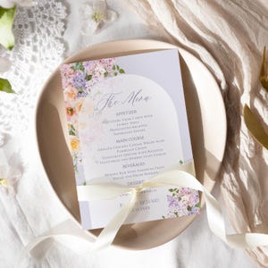 May include: A purple and white floral wedding menu card with a white ribbon tied around it. The menu includes appetizers, main course, beverages, and dessert. The text on the card reads "The Menu", "Appetizer", "Miniature Crab Cakes with Lemon Aioli", "Prosciutto-Wrapped Asparagus Spears", "Main Course", "Grilled Chicken Caesar Wraps with Parmesan Crisps", "Vegetarian Quiche with Roasted Vegetables and Herbed Goat Cheese", "Beverages", "Mimosa Bar with Assorted Fruit Juices and Fresh Berries", "Dessert", "With Sprinkles", "JPCHARD", "2.12 20XX".