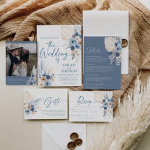 May include: A wedding invitation suite with a blue and beige color scheme. The invitation features a floral design with pampas grass and the text "The Wedding of Sarah and Thomas". The details card includes information about the wedding reception, directions, and accommodations. The RSVP card has a space for guests to provide their names and indicate whether they will be attending. The gifts card includes a message about the couple's registry.