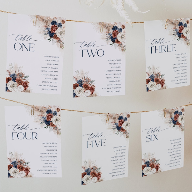 May include: Six white rectangular wedding seating charts hanging on a twine string with gold clips. Each card features navy blue text, floral arrangements in navy, rust, and cream, and the words Table One through Table Six.