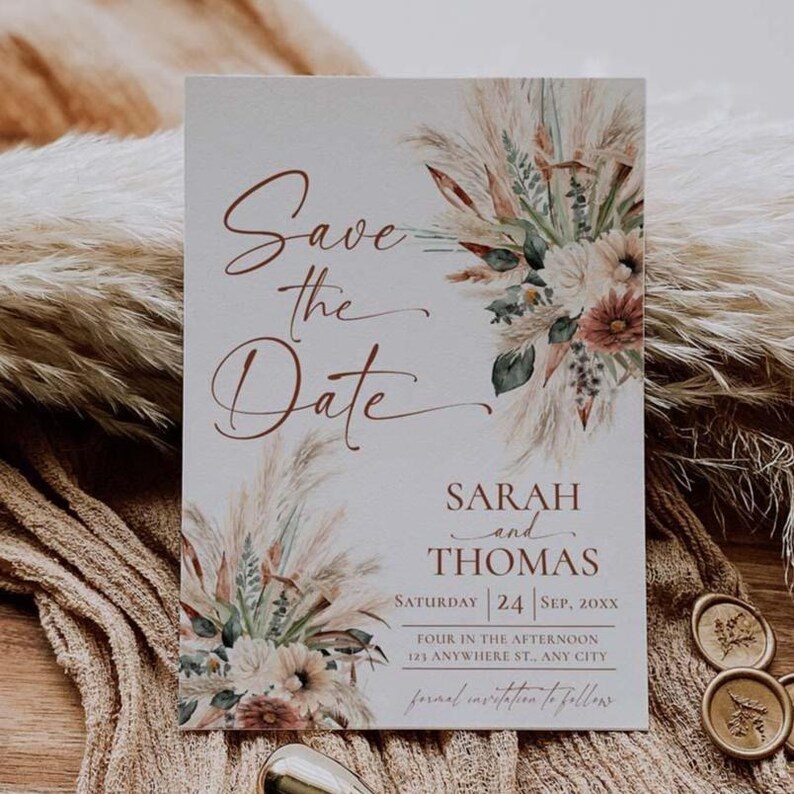 May include: A rectangular Save the Date card featuring copper-coloured cursive text and printed illustrations of pampas grass and dried flowers. The card displays the names Sarah and Thomas, the date Saturday 24 Sep, 20XX, and event details.