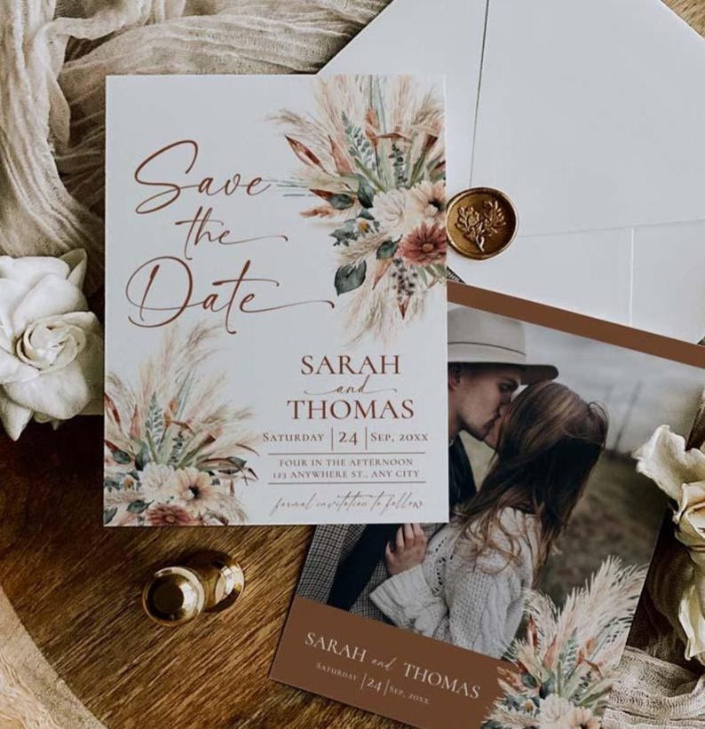 May include: A Save the Date card featuring pampas grass and floral illustrations, with the text Sarah and Thomas, Saturday 24 Sep, 20xx. Includes a photo card of a couple, a gold wax seal, and a white envelope on a wooden surface.