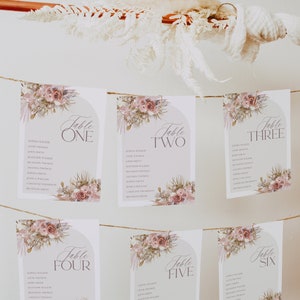May include: Six floral table cards with a light pink and brown floral design. Each card has a table number, from one to six, and a list of names. The cards are strung together with twine.