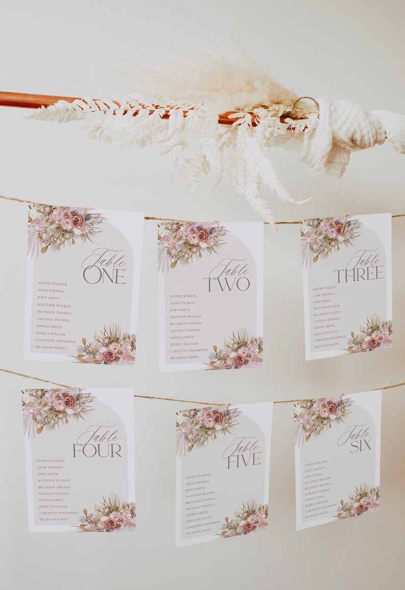 May include: Six floral table cards with a light pink and brown floral design. Each card has a table number, from one to six, and a list of names. The cards are strung together with twine.